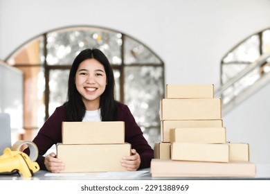 Female young online business, store owner or entrepreneur sitting at desk with cardboard boxes, scanner, and tape holding parcel and looking at camera with happy and smiley face.  - Powered by Shutterstock