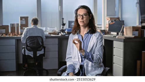 Female young scientist in lab coat talks to the camera and gives interview for popular science film while sitting in archaeological scientific lab. Male colleague works in the background. Static Shot. - Powered by Shutterstock