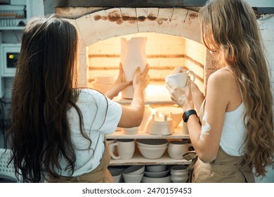 Females ceramic artisans arranging clay mugs in kiln at pottery workshop - Powered by Shutterstock