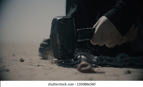 Female's Hand Hitting A Nail With A Hammer In Desert. Chains And Hammer In Desert. Slavery In Dune. Dark Silhouette On The Back. Dust And Particles. Freeze Frame.Cinematic Shot. Cold Color Correction.