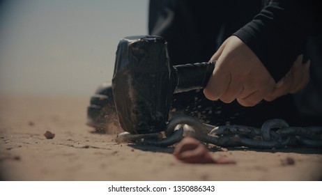 Female's Hand Hitting A Nail With A Hammer In Desert. Chains And Hammer In Desert. Slavery In Dune. Dark Silhouette On The Back. Dust And Particles. Freeze Frame.Cinematic Shot. Warm Color Correction.