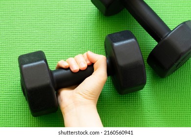 Female's Hand Holding A Dumbbell On Green Workout Mat Background. Top View, Flat Lay. Strong Woman, Lifting Weights, Exercise At Home Concept. A Close-up.