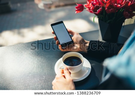 Female's hand holding mobile phone with empty white screen next to cup of coffee at table in cafe by window, close-up.