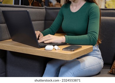 Female`s hands  with cell phone in front of laptop computer, sits at cafeteria, surrounded with mug of coffee and modern laptop and wireless headphones on table