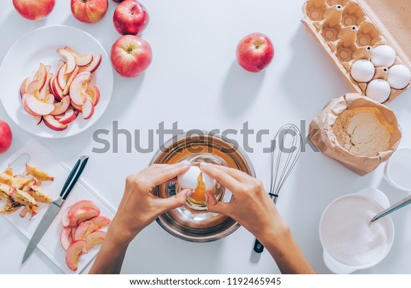 Female\'s hands cracking egg into bowl divide its\
shell next to ingredients for baking apple pie, top view. Flat\
layout young woman prepares dough for cake surrounded cut fruit,\
flour and sugar.