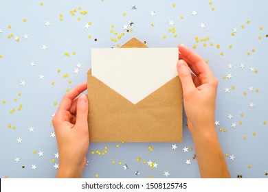 Female's Hands Holding Kraft Paper Envelope Letter With Blank White Card Mockup Over Pastel Blue Background With Golden Confetti Stars. Christmas, New Year, Winter Holidays And Birthday Concept.