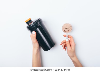 Female's Hands Holding Scoop Of Chocolate Protein Powder And Black Plastic Shaker To Prepare Sports Drink For Muscle Growth, Top View On White Table.