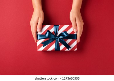 Female's Hands Holding Striped Gift Box With Blue Ribbon On Red Background. Christmas, New Year, Valentine's Day And Birthday Concept.