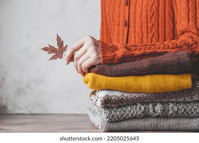 A Females's Hand Holding A Fallen Leaf Leaning On Stack Of Knitted Sweaters Autumn Color Palette. Fashion And Sustainability Life. High Quality Photo