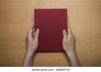 A Female(woman) Hands Hold A Blank(empty) Red Book(note, Diary) Cover On The Wood Desk(table), Top View At The Studio.