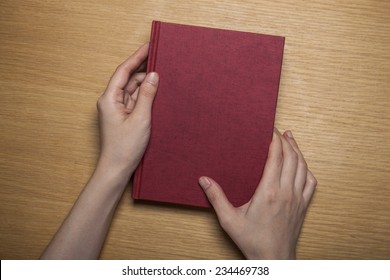 A Female(woman) Hands Hold A Blank(empty) Red Book(note, Diary) Cover On The Wood Desk(table), Top View At The Studio.