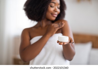 Feminine black woman holding jar of body or face cream, applying skin care product after shower at home, selective focus. Sensual Afro lady using cosmetics in morning, indoors
