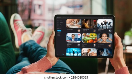 Feminine Hand Holding A Tablet With Video Streaming Service On Display. Female Is Relaxing On A Couch At Home, Watching Videos And Reading Social Media Posts On Mobile Device. Close Up POV Photo.