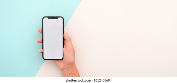 Feminine Hand Holds IPhone X With White Empty Screen. Wide Mockup For Billboard Or Screenshot App With Space For Features