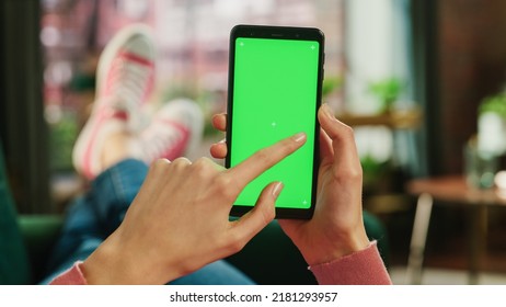 Feminine Hand Scrolling Feed On Smartphone With Green Screen Mock Up Display. Female Is Relaxing On Sofa At Home, Watching Videos And Reading Social Media Posts On Mobile Device. Close Up POV Photo.