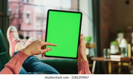 Feminine Hand Scrolling Feed On Tablet With Green Screen Mock Up Display. Female Is Relaxing On Sofa At Home, Watching Videos And Reading Social Media Posts On Mobile Device. Close Up POV Photo.