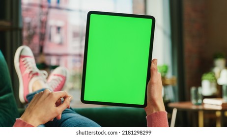 Feminine Hand Scrolling Feed On Tablet With Green Screen Mock Up Display. Female Is Relaxing On Sofa At Home, Watching Videos And Reading Social Media Posts On Mobile Device. Close Up POV Photo.