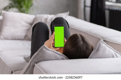 Feminine Hand Scrolling Feed on Smartphone with Green Screen Mock Up Display. Female is Relaxing on Sofa at Home, Watching Videos and Reading Social Media Posts on Mobile Device. - Shutterstock ID 2258236097