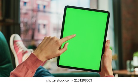 Feminine Hand Swiping Feed On Tablet With Green Screen Mock Up Display. Female Is Relaxing On Sofa At Home, Watching Videos And Reading Social Media Posts On Mobile Device. Close Up POV Photo.