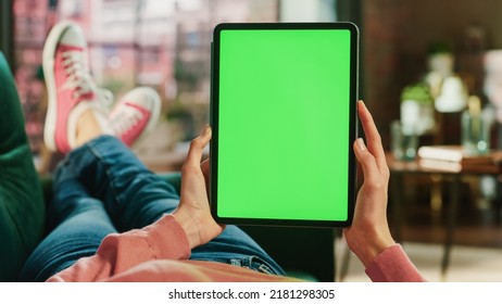 Feminine Hand Vertically Holding A Tablet With Green Screen Display. Female Is Relaxing On A Couch At Home, Watching Videos And Reading Social Media Posts On Mobile Device. Close Up POV Photo.