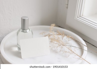Feminine lifestyle cosmetic still life scene. Glass bottle, flacon with perfume or eau de toilette and dry grass on white marble tray near window. Blank business card mockup. Selective focus.