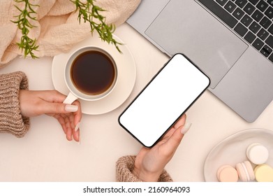 Feminine office desk with female hand using smartphone and sipping coffee. mobile phone blank screen mockup. top view