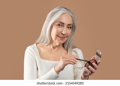 Feminine Senior Woman Holding Cosmetic Brush And Eye Shadow Or Blusher Palette On Brown Studio Background. Beautiful Mature Lady Using Decorative Makeup, Pampering Herself