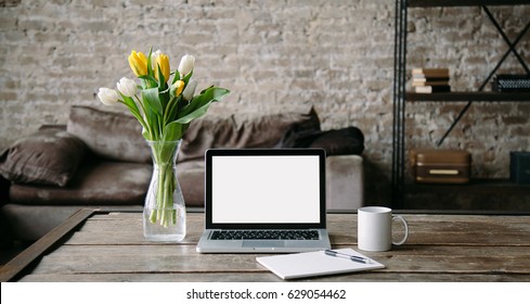 Feminine workplace front view with laptop screen mock up, coffee mug, bouquet of yellow tulips and empty page with pencil. Creative concept. Blogger's decorated workspace.