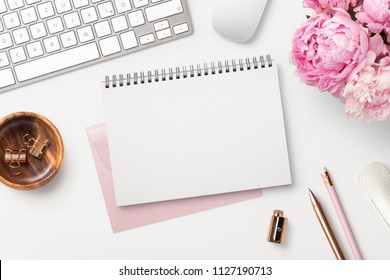 feminine workspace / desk with blank open notepad, keyboard, stylish office / writing supplies and pink peonies on a white background, top view