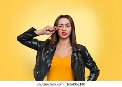 Feminism And The Concept Of Equal Rights And Independence For Women. A Caucasian Young Woman In A Leather Jacket Draws War Paint On Her Face With Lipstick. Isolate. Yellow Background. Copy