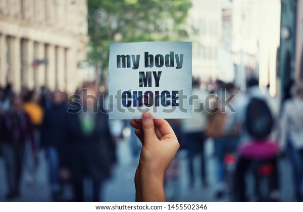 Feminist hands holding a protest banner with\
the message my body my choice over a crowded street. Human rights\
concept against fetus law and reproductive justice. Stop\
discrimination and\
injustice.