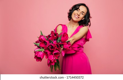 Feminity. Close-up photo of a girl in a fuchsia dress, who is holding a bunch of pink tulips in her stretched hands, looking in the camera with joy. - Powered by Shutterstock