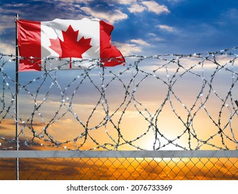 A Fence With Barbed Wire Against The Background Of A Colorful Sky And A Flagpole With The Flag Of Canada Protects The State's Border From Illegal Migration.