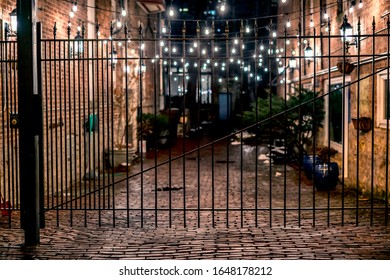 A fence in front of a string lit brick vintage alley way brightened with yellow string lights at night in an urban neighborhood.