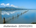 Fence leading into calm blue water of Lake Wairarapa with distant hills across other side in New Zealand North Island.