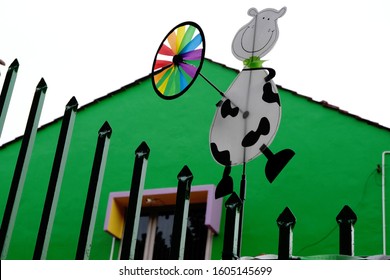 A Fence Outside A Nursery Decorated With Children's Cartoon Animal Characters. A Plastic Cutout Of A Cow Holding A Pinwheel Lines The Fencing Of A Childcare Center. Barrier Gate Of A Daycare Building.