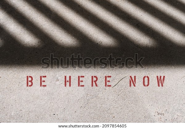 Fence shadow on\
cement floor with text inscription BE HERE NOW, concept of live\
with present, power of living with now - be with the moment, not\
worry about past or\
future