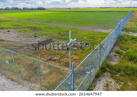 Fence surrounds a field of grass. A camera is mounted on a pole in the middle of the field
