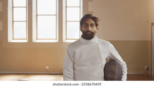 Fencer in white uniform holding helmet, standing confidently in bright room. Fencing, athlete, sword, competition, sportsman, confidence - Powered by Shutterstock