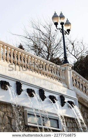 Fencing on an antique white stone marble fence in city park with a cascade of artificial streams of water, waterfalls, fountains. Metal black street lamp Vertical background with architectural details