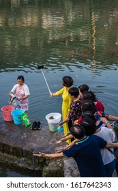 Feng Huang, China - August 2019 : Chinese Family Taking Selfie Photo On The Riverbanks Of Tuo River, While Older Woman Washes Laundry And Clothes On The Shore