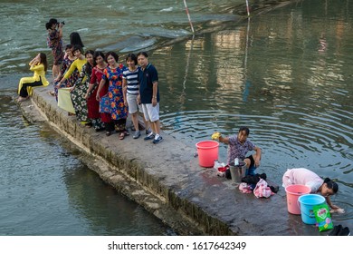 Feng Huang, China - August 2019 : Chinese Family Taking Selfie Photo On The Riverbanks Of Tuo River, While Older Woman Washes Laundry And Clothes On The Shore