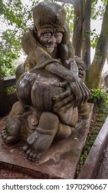 Fengdu, China - May 8, 2010: Ghost City, Historic Sanctuary. Brown Stone Statue Of Old Man With Sharp Teeth Handling Naked Behind Of Young Woman Who Seems To Enjoy It. Green Foliage.