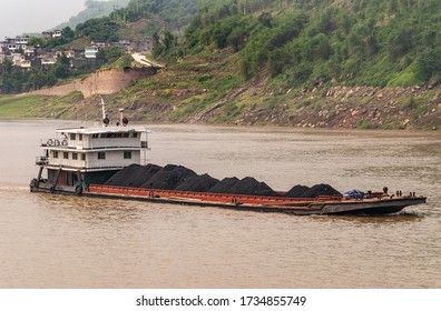 Fengdu, Chongqing, China - May 8, 2010: Yangtze River. Closeup Of Barge Filled With Heaps Of Black Coal On Brown Water With Green Shoreline And Some Residential Housing.