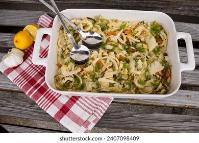 Fennel with orange, capers and parmesan in a white dish fresh from the oven on red checkered cloth. Serving cutlery, lemon and garlic bulb, served on a wooden bench - Powered by Shutterstock