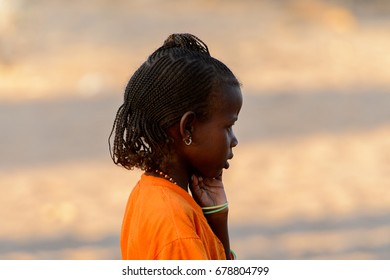 FERLO DESERT, SENEGAL - APR 25, 2017: Unidentified Fulani Little Girl With Braids In Orange Shirt Stands On The Street. Fulanis (Peul) Are The Largest Tribe In West African Savannahs