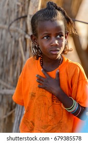 FERLO DESERT, SENEGAL - APR 25, 2017: Unidentified Fulani Little Girl With Braids In Orange Shirt Looks Away. Fulanis (Peul) Are The Largest Tribe In West African Savannahs