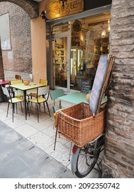 Ferrara, Italy - December 4, 2021. Bistro In The Medieval Area. Outside There Are Colorful Tables And Chairs. A Blackboard With The Menu Rests On The Wicker Basket Of An Old Bike.