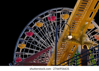 Ferris wheel and roller coaster structure at Santa Monica Pier at night with palm tree and pedestrian silhouette, ideal for travel, nightlife, and coastal lifestyle concepts.