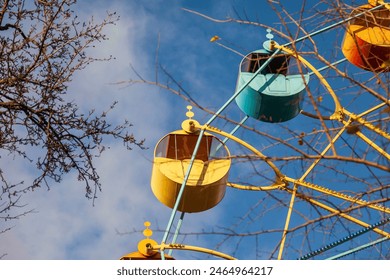 A Ferris wheel with yellow and blue booths for tourist passengers floating across a blue cloudy sky. Blurred background of trees with spring swelling buds. - Powered by Shutterstock